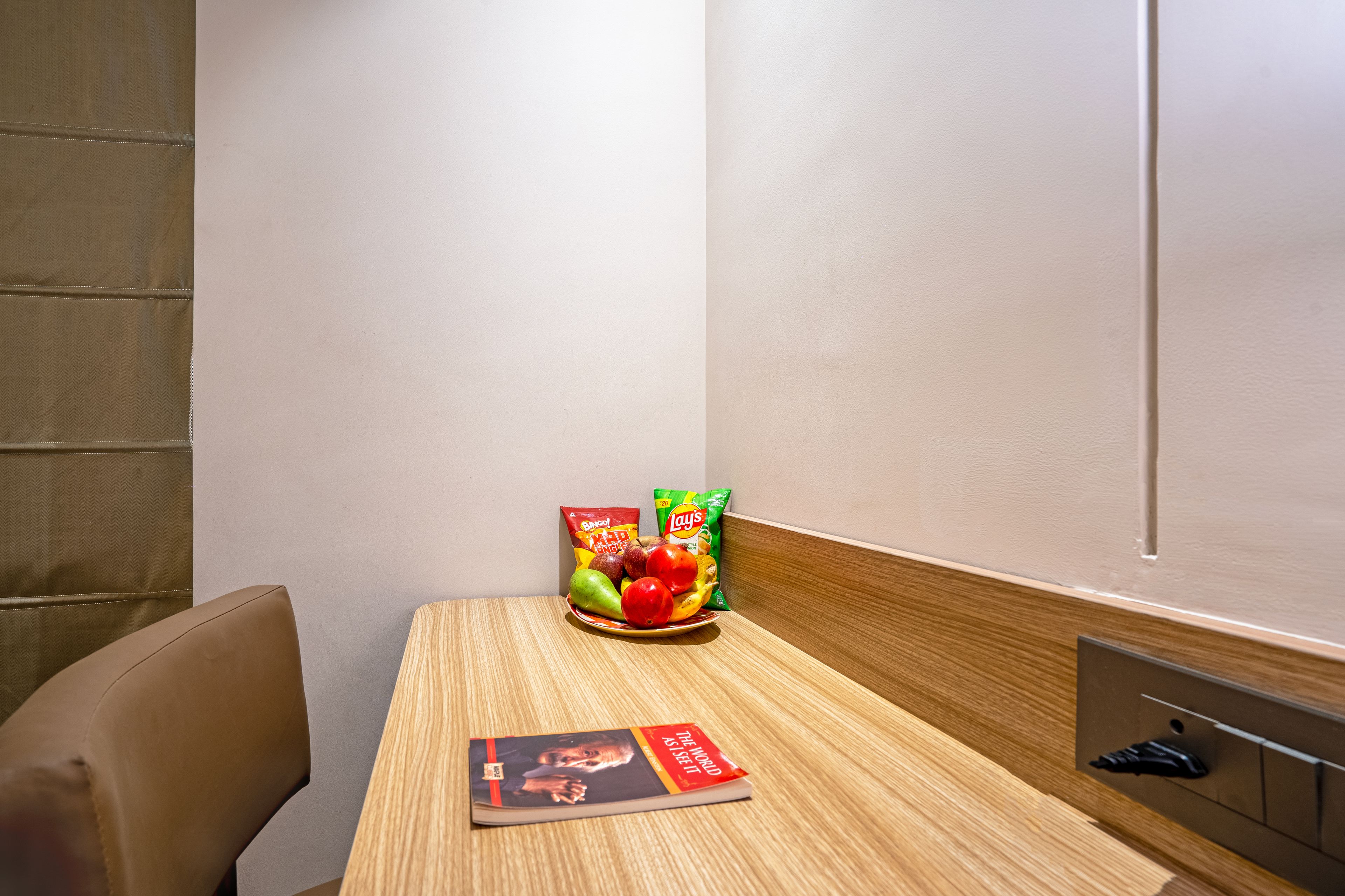 A hotel room with a wooden table, a magazine, a plate of fruit, and snack packets.