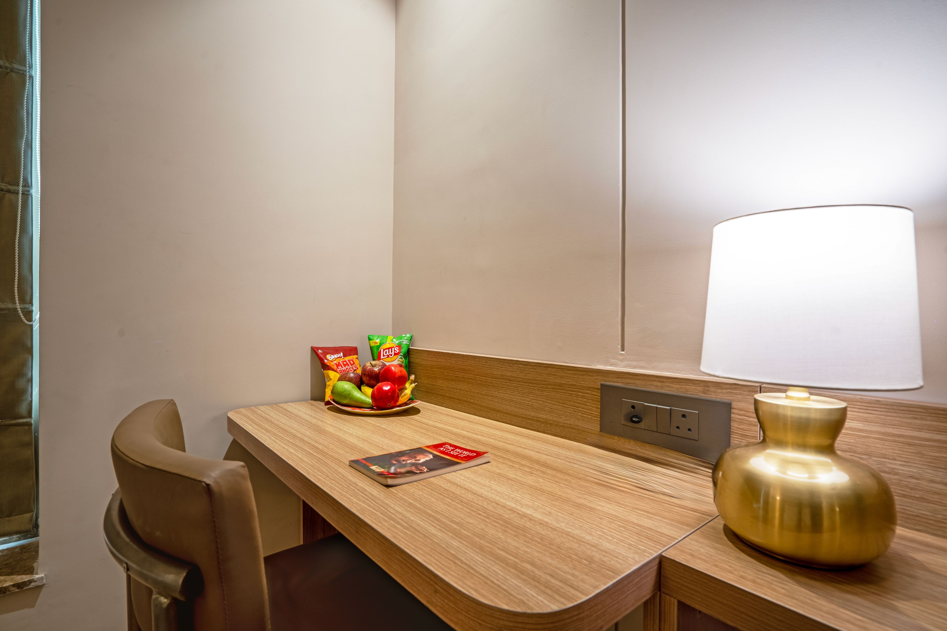 Habitación de hotel con un escritorio de madera, una lámpara, una silla y un frutero con aperitivos.