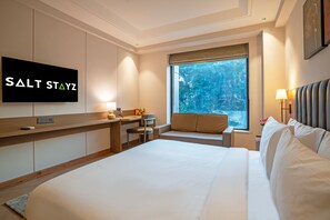 A hotel room with a bed, a desk, a chair, a television, and a window with a view of trees.