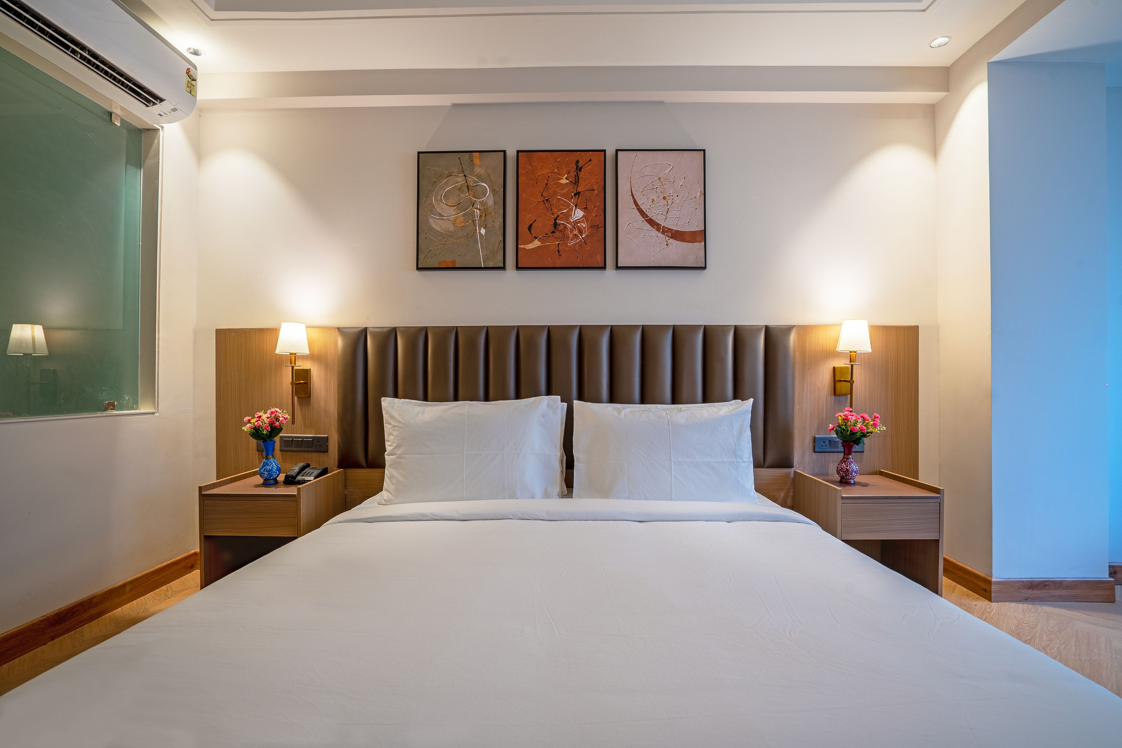 A hotel room with a large bed, two bedside tables with lamps, a wall-mounted air conditioner, and three framed artworks above the bed.