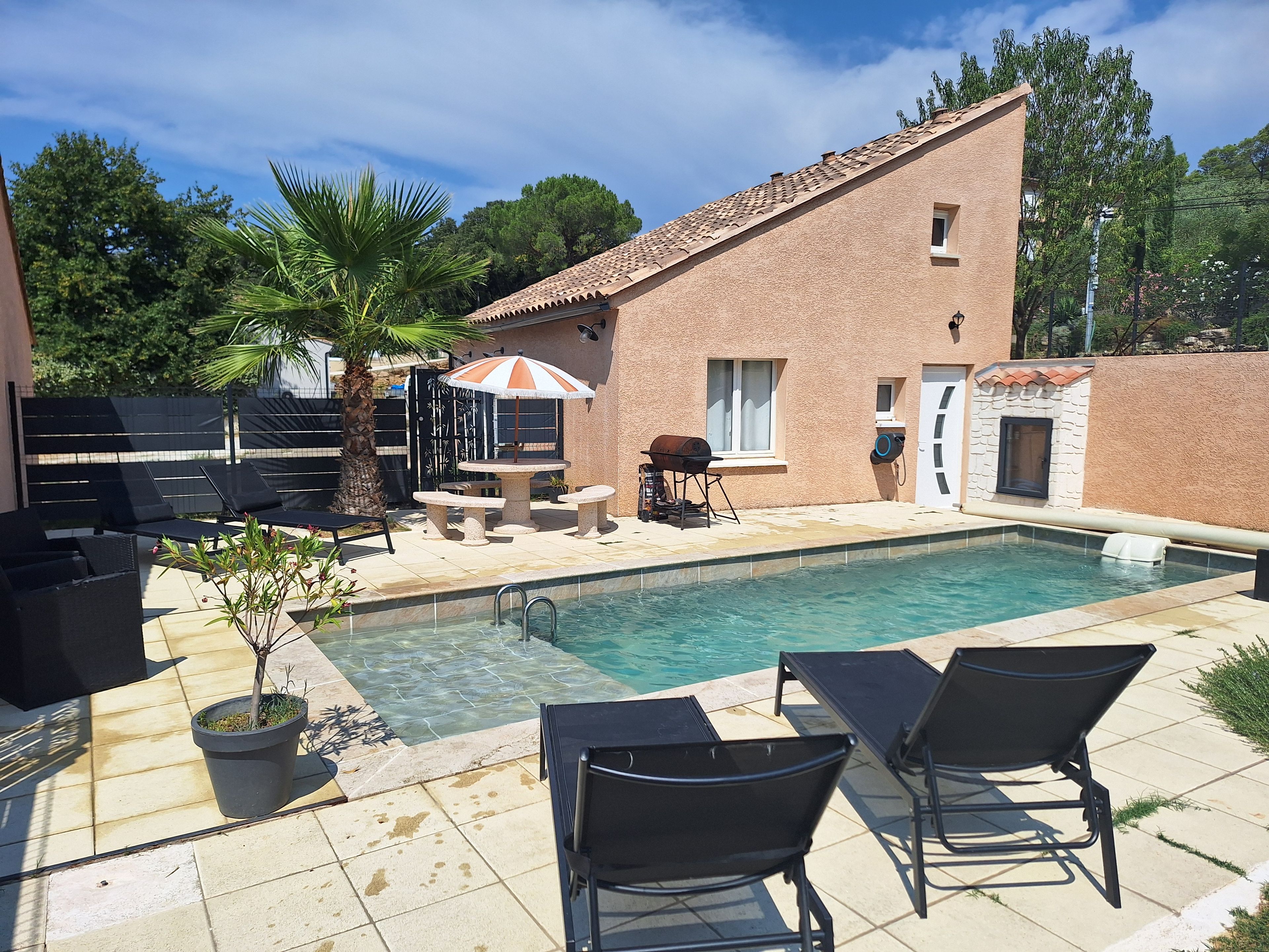 Une piscine avec des chaises longues et un espace barbecue, le tout entouré d’un bâtiment en briques et de palmiers.