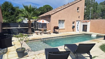 Une piscine avec des chaises longues et un espace barbecue, le tout entouré d’un bâtiment en briques et de palmiers.