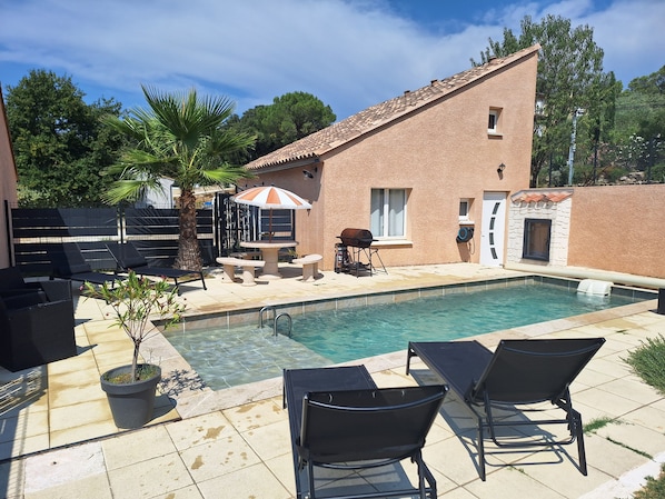 Une piscine avec des chaises longues et un espace barbecue, le tout entouré d’un bâtiment en briques et de palmiers.