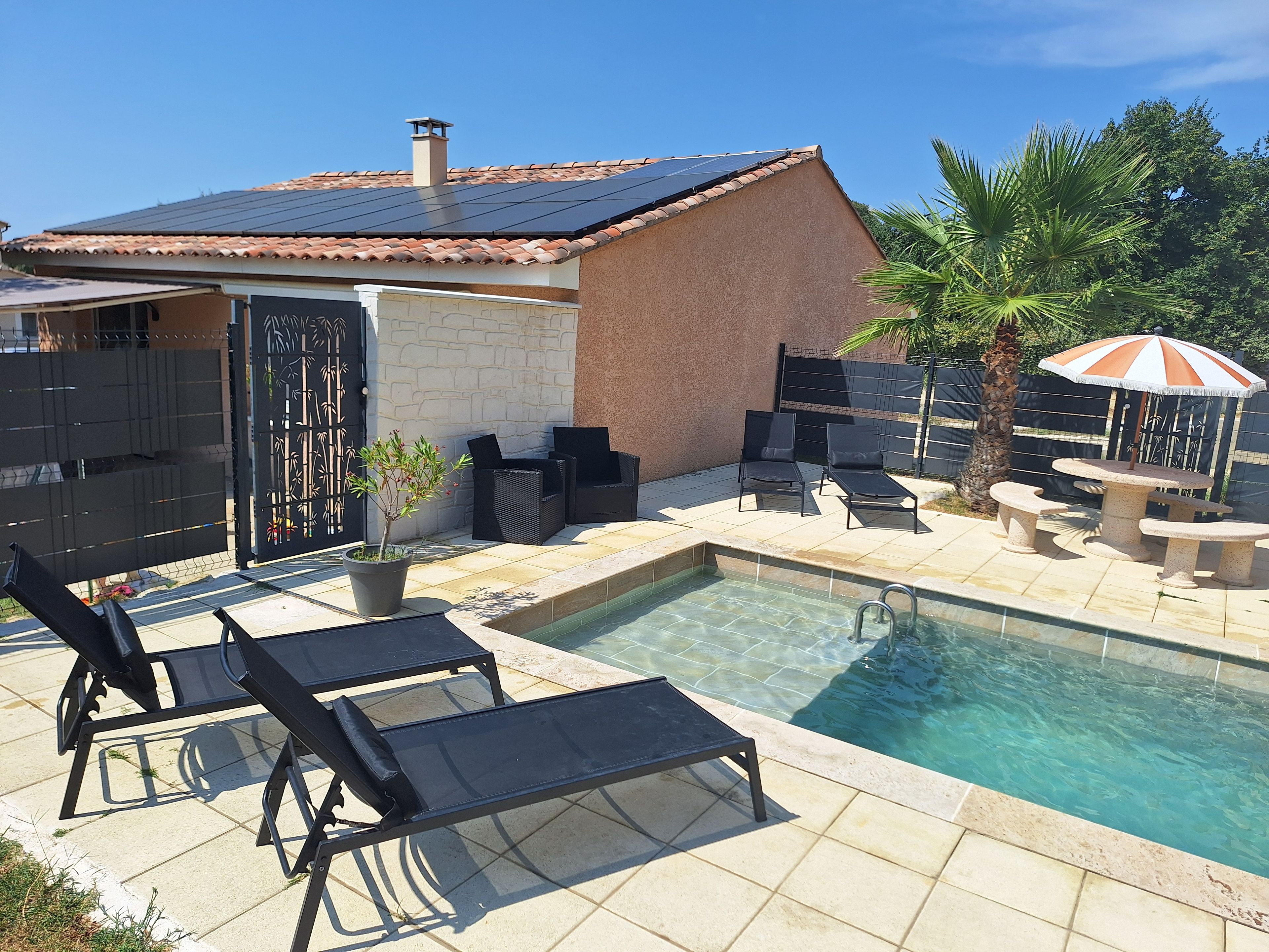 Une piscine entourée de chaises longues et d’une table, le tout délimité par une terrasse clôturée agrémentée d’un palmier et d’un parasol décoratif.