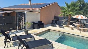 Une piscine entourée de chaises longues et d’une table, le tout délimité par une terrasse clôturée agrémentée d’un palmier et d’un parasol décoratif.