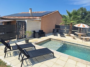 Une piscine entourée de chaises longues et d’une table, le tout délimité par une terrasse clôturée agrémentée d’un palmier et d’un parasol décoratif.