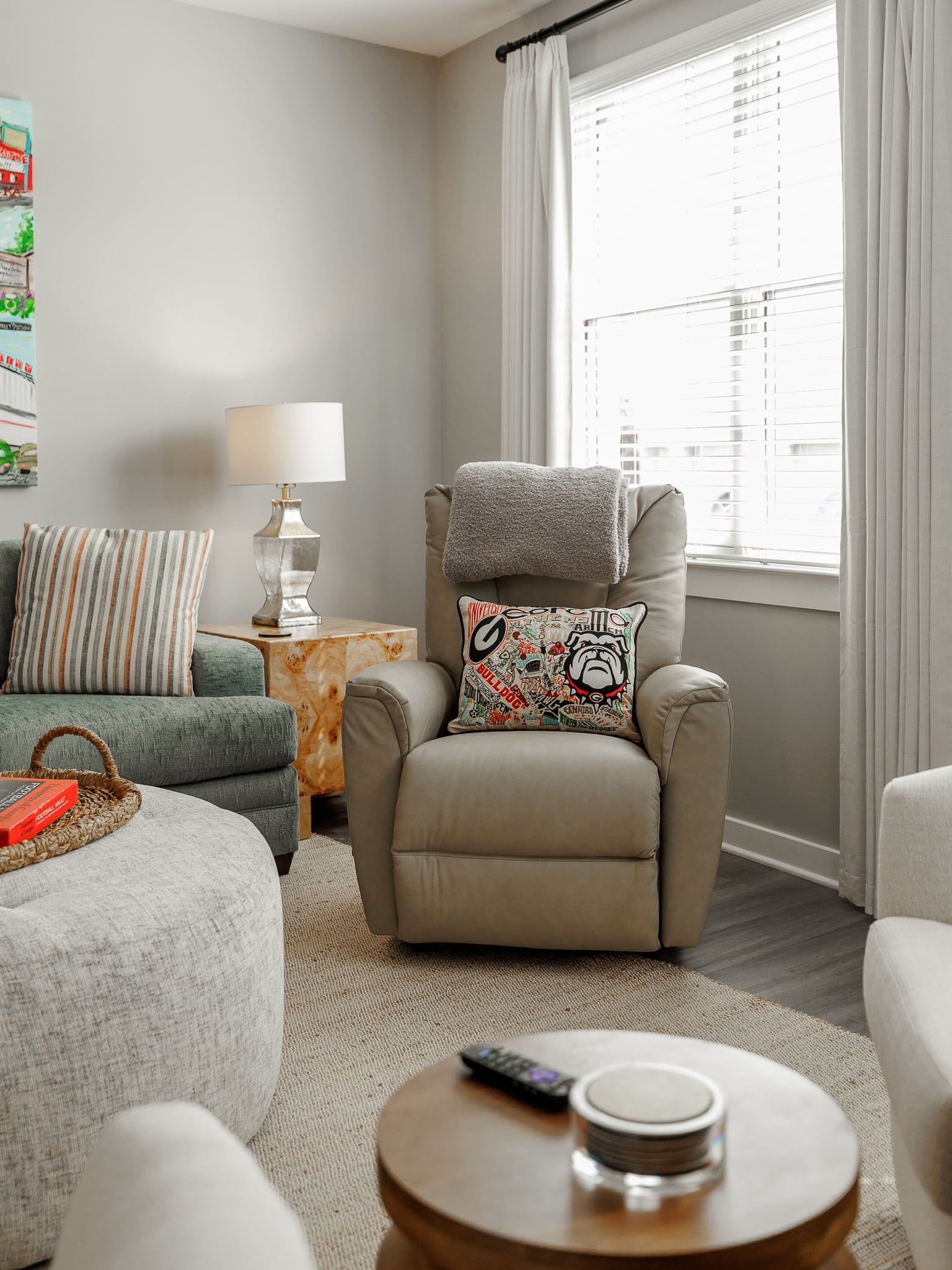 Een zithoek met een beige fauteuil, een grijze plaid, een kussen met dessin, een klein bijzettafeltje met een lamp en een afstandsbediening op een ronde salontafel.