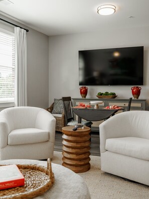 A modern living room with two white armchairs, a round wooden coffee table, a flat-screen TV mounted on the wall, and a dining table with red vases and a bowl of green vegetables. - The Athens Social - walk to downtown! (Athens)