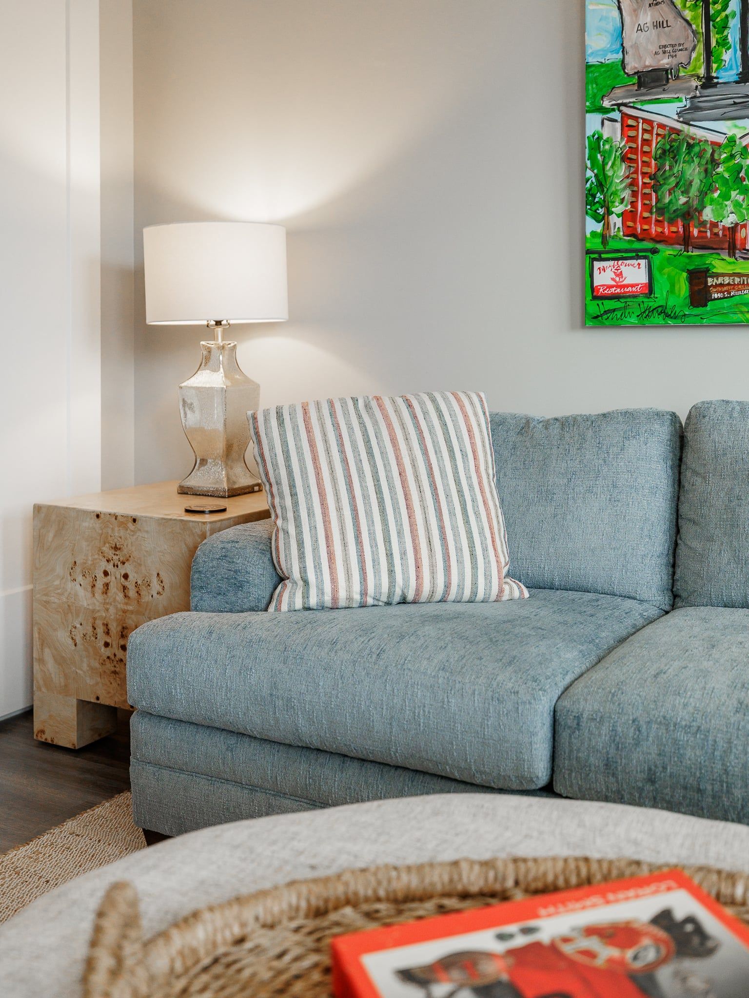 A modern living room with a grey sofa, a striped pillow, a lamp on a wooden side table, and a framed painting on the wall.