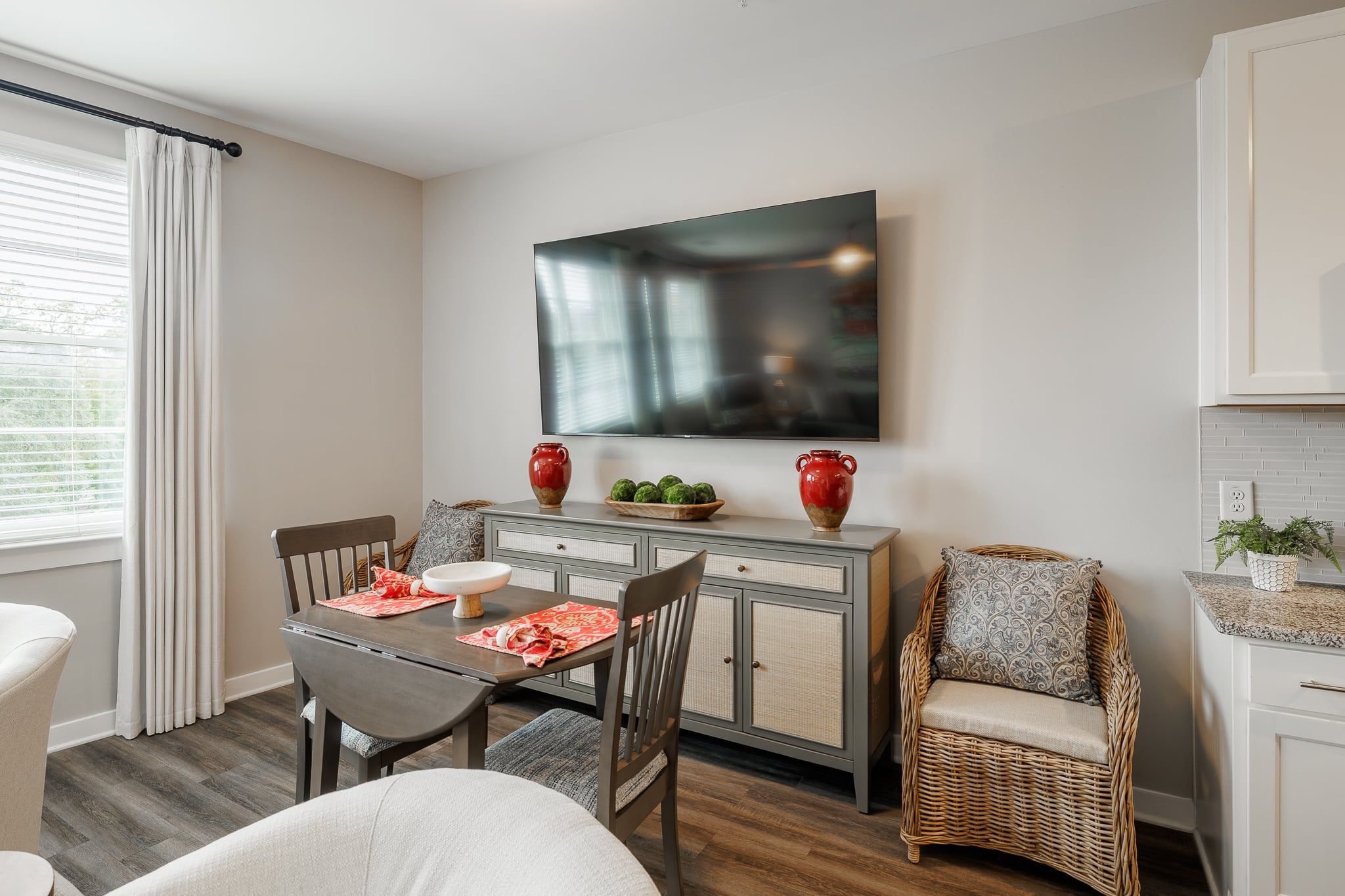 A dining area with a table, chairs, a television mounted above, and a wicker chair.