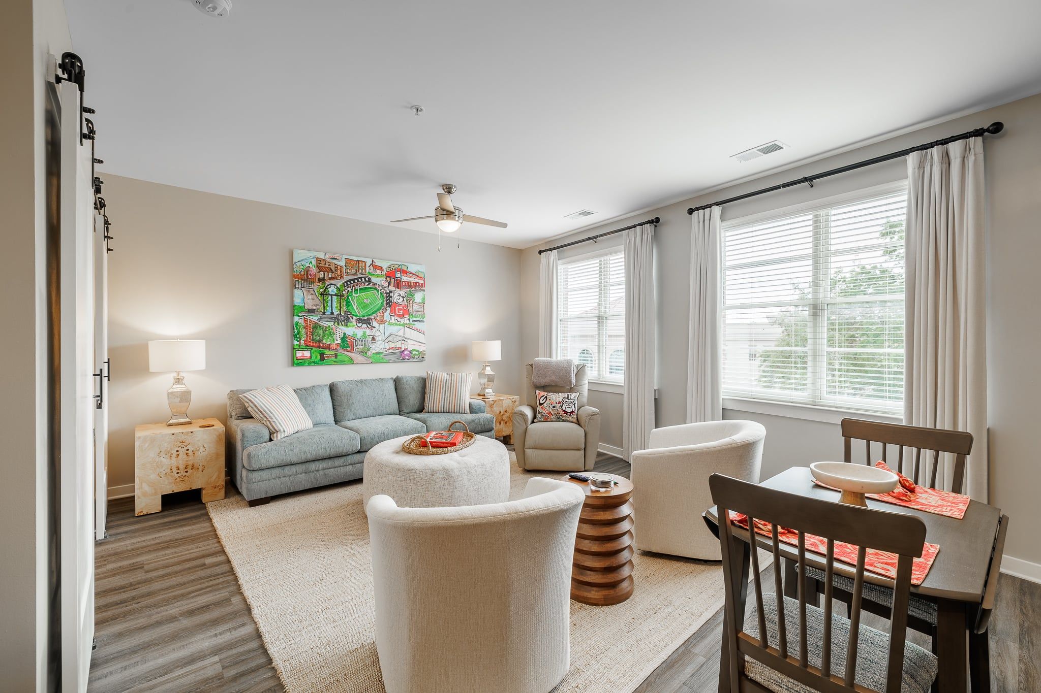 A living room with a sofa, ottoman, chairs, a coffee table, a painting, a ceiling fan, and a window with curtains.