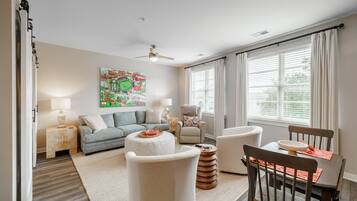 A living room with a sofa, ottoman, chairs, a coffee table, a painting, a ceiling fan, and a window with curtains.