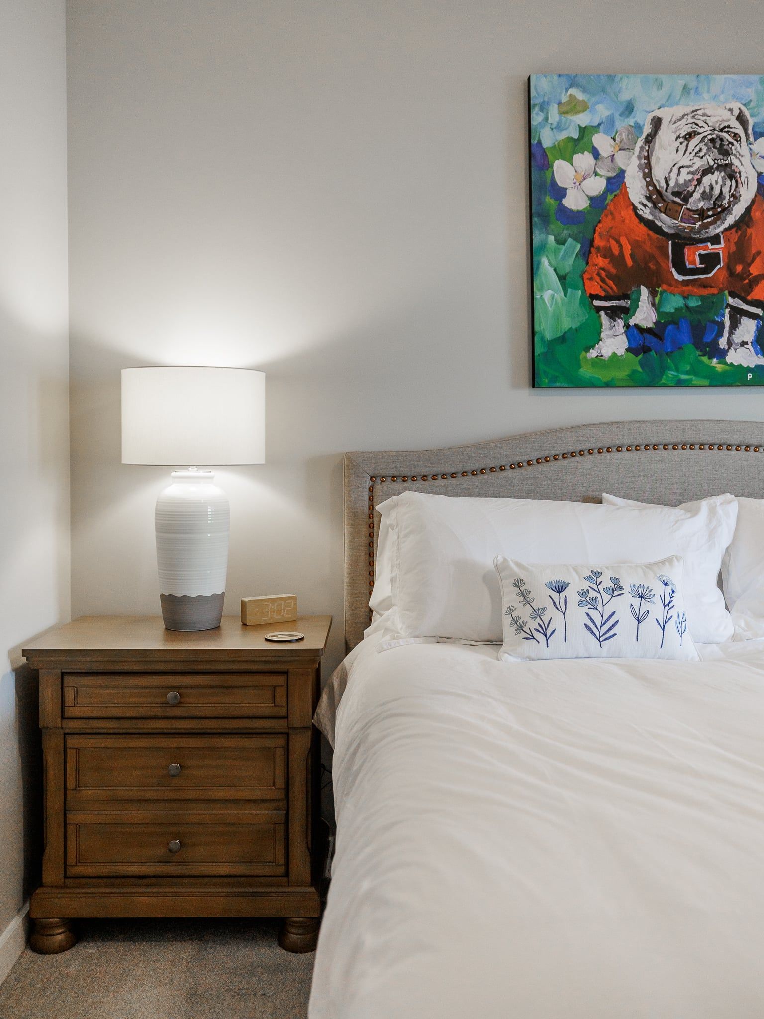 Ein Schlafzimmer mit einem Bett, weißer Bettwäsche, einem Nachttisch mit einer Lampe, einer Wasserflasche, einer digitalen Uhr und einem Gemälde eines Bulldoggen an der Wand.