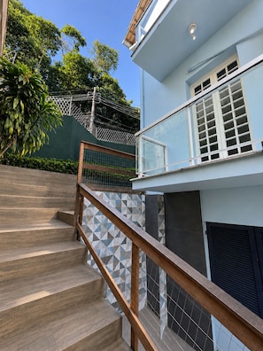 A staircase with wooden steps and a metal railing leading to a modern building with a light blue exterior and a balcony. - A different Rio experience | Exclusive house with 4 suites near Barra. (Rio de Janeiro)