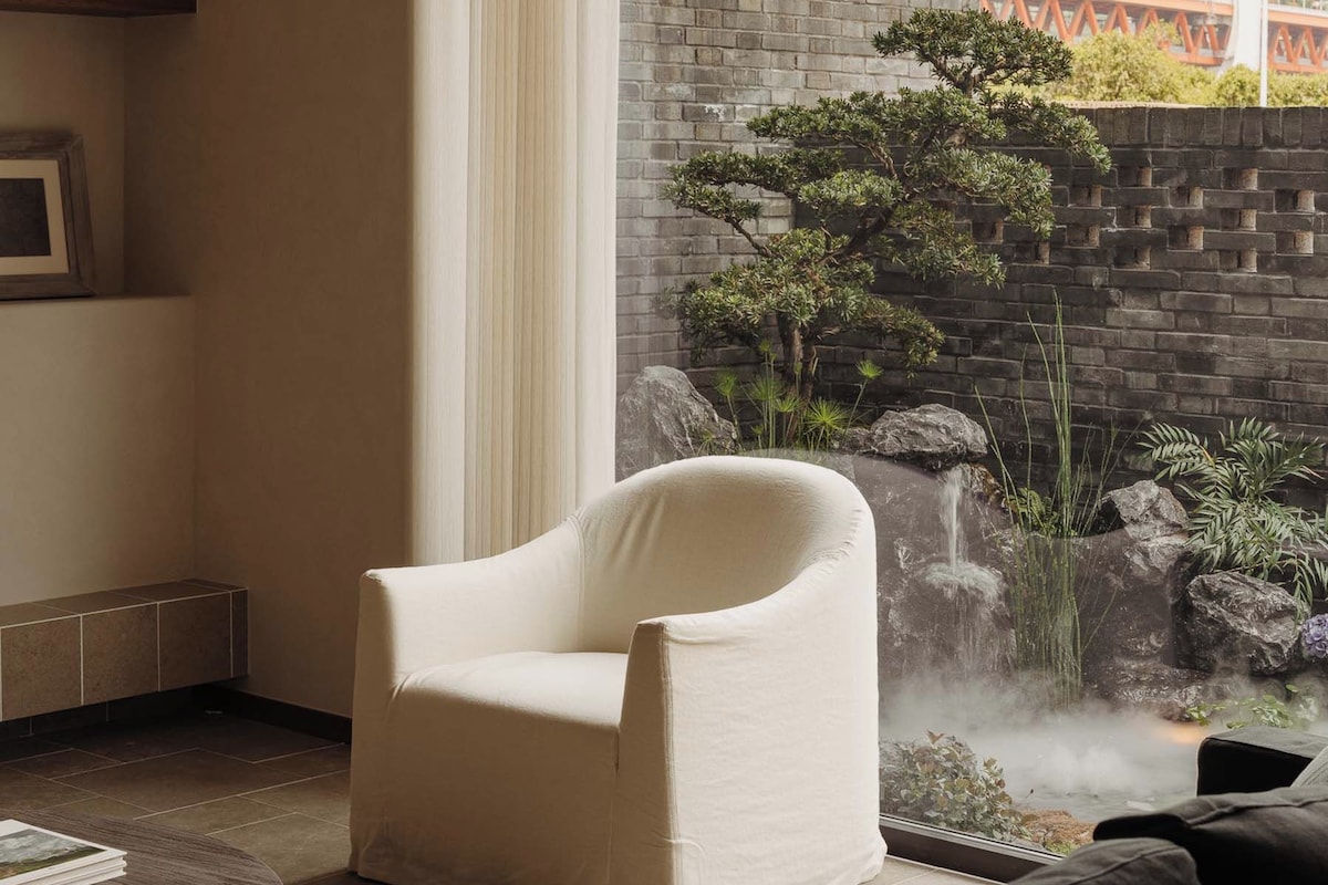 Un salon moderne avec un fauteuil blanc, une table basse ronde et une vue sur un pont et la verdure environnante.