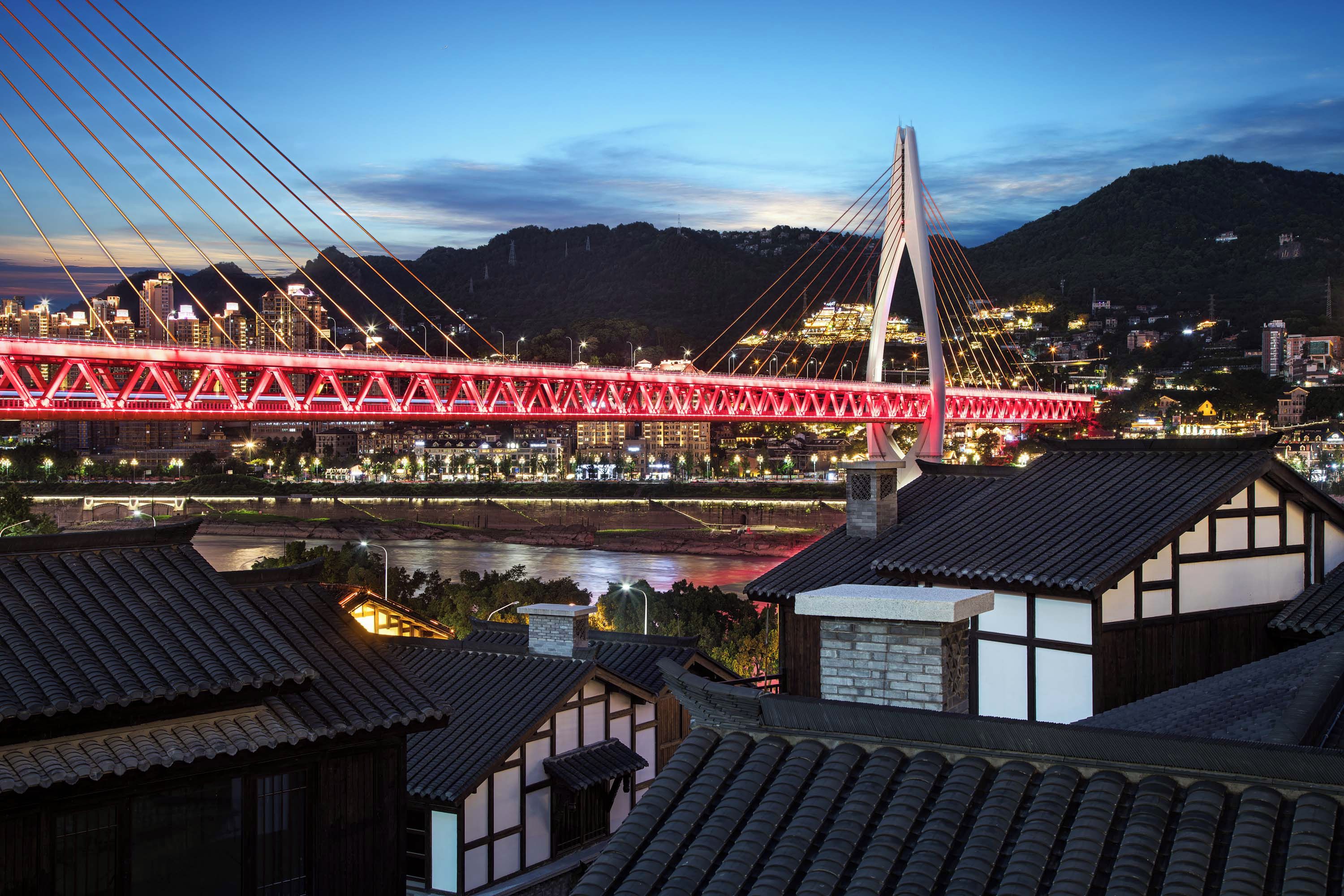 Un moderno puente atirantado con iluminación roja, un río debajo y edificios tradicionales al frente.