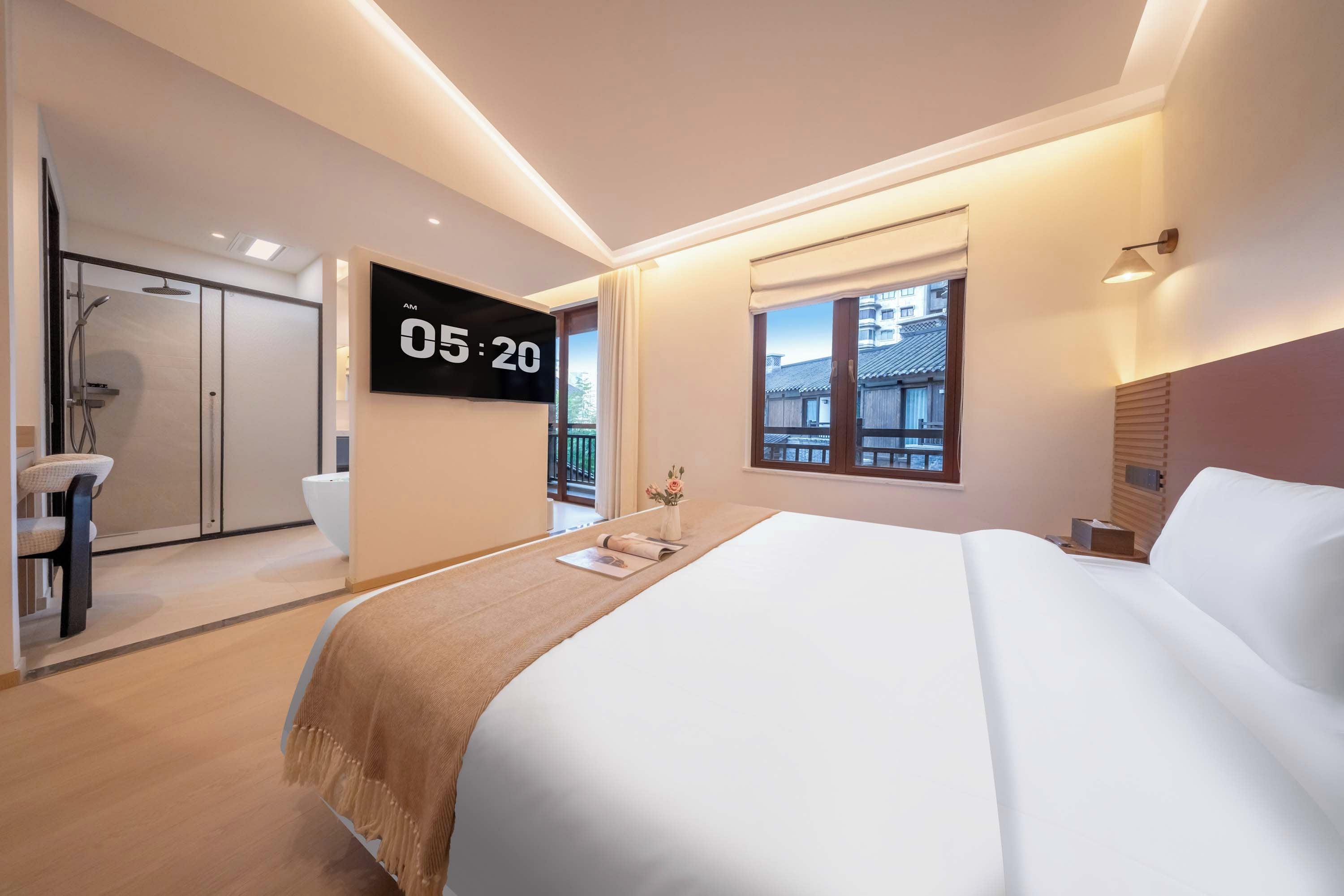 A modern hotel room with a bed, a TV displaying the time, a window with a view of buildings, and a shower area.