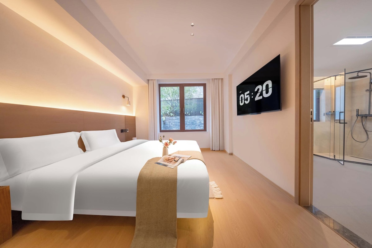 Une chambre d’hôtel moderne avec un grand lit, une table de chevet avec un magazine, une horloge numérique et une cabine de douche en verre.