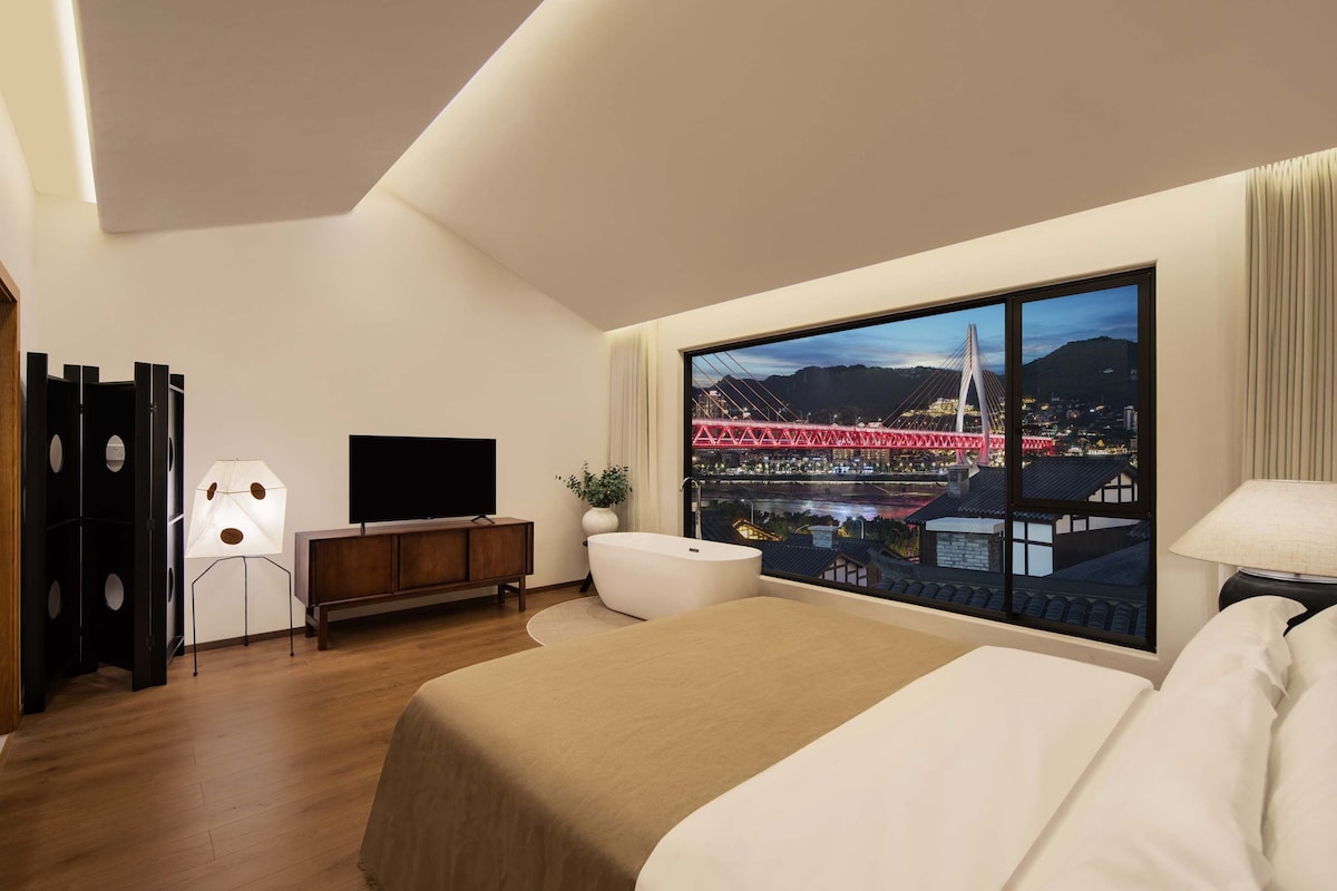 Une chambre d’hôtel moderne dotée d’une grande fenêtre, d’un téléviseur à écran plat, d’une armoire en bois, d’une baignoire blanche et d’un lit recouvert d’une courtepointe beige.