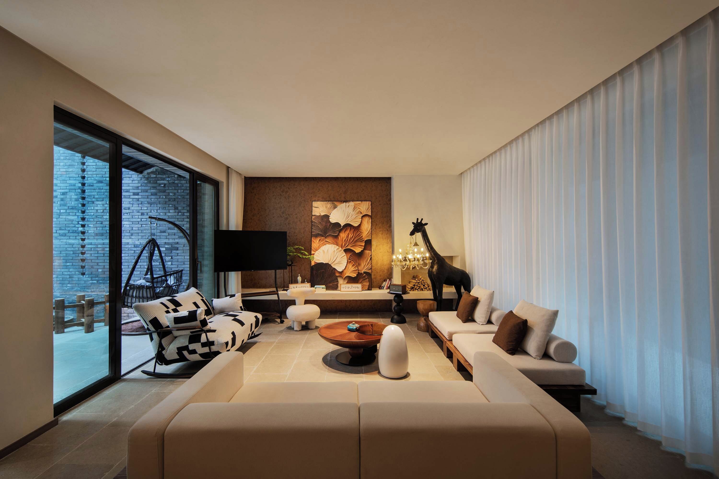 Una sala de estar moderna con un sofá grande, una mesa de centro y un televisor. Hay un ventanal con vista a un muro de ladrillo y una silla colgante afuera.