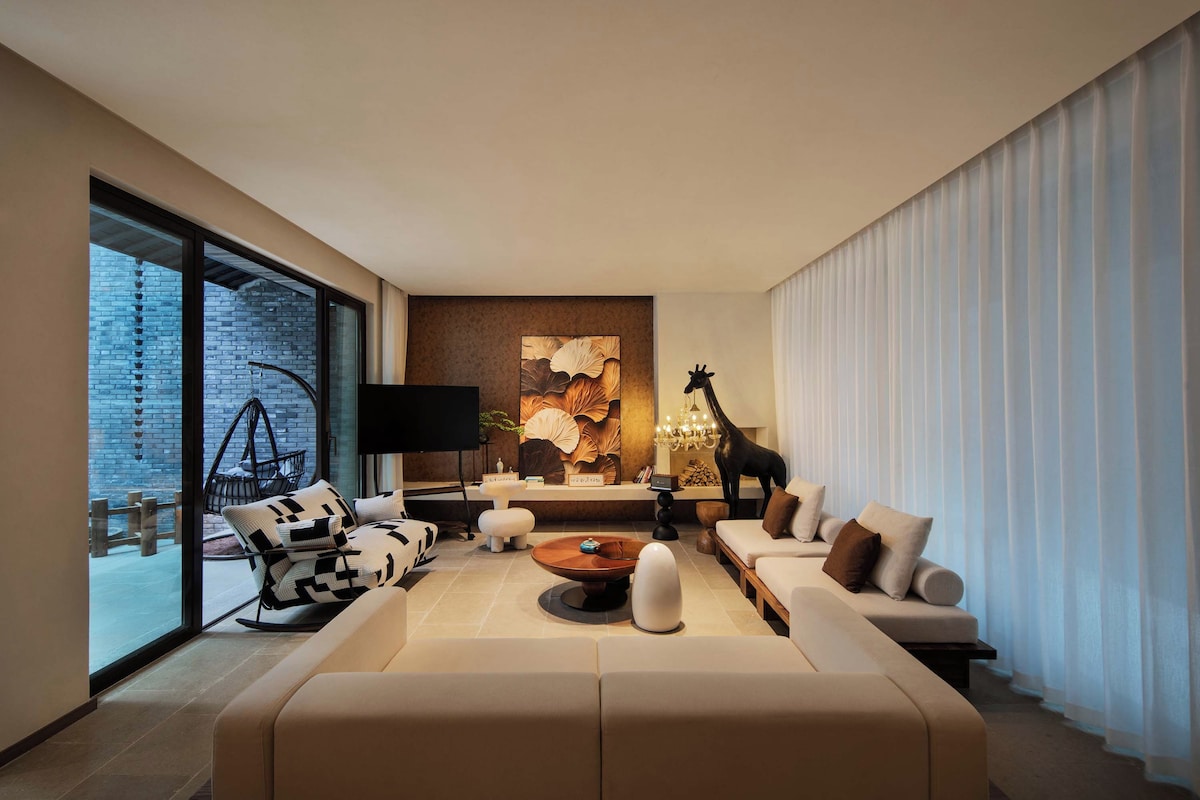 Un salon moderne avec un grand canapé, une table basse et un téléviseur. Une grande fenêtre offre une vue sur un mur de briques et on aperçoit une chaise suspendue à l’extérieur.