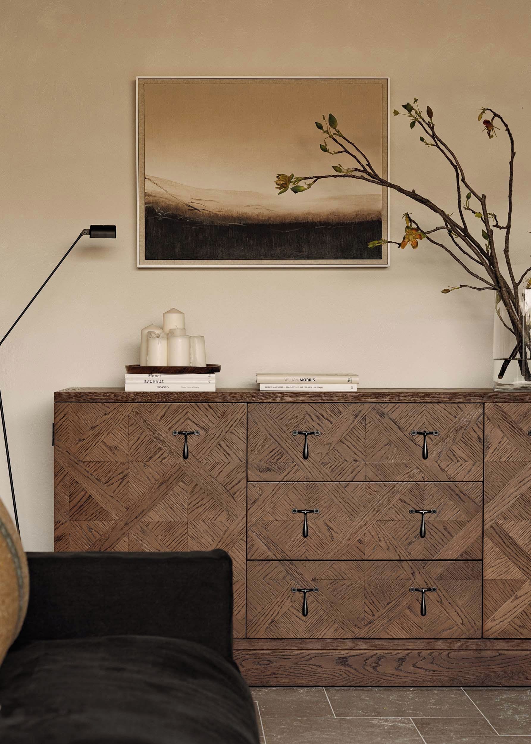 Una habitación con un armario de madera con motivo de espiga, un cuadro enmarco de un paisaje, y un jarrón con ramas y flores sobre una mesa.