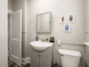 Ein Badezimmer mit weißem Waschbecken, Spiegel und Toilette. An der Wand hängen drei Bilder in Rahmen.