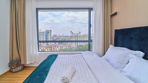 A hotel room with a large window, a bed with white linens, a dark headboard, and a view of city buildings. - Charming apartment in beautiful Kilimani, Nairobi perfect for your stay (Nairobi)