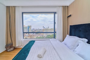 A hotel room with a large window, a bed with white linens, a dark headboard, and a view of city buildings. - Charming apartment in beautiful Kilimani, Nairobi perfect for your stay (Nairobi)