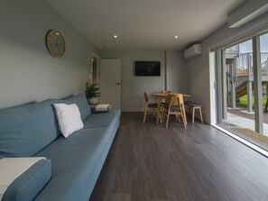 A living room with a blue sofa, a wooden dining table with chairs, a TV mounted on the wall, and a large window.