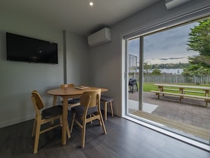 A room with a dining table and chairs, a flat-screen TV mounted on the wall, and a large sliding glass door leading to an outdoor patio with a bench.