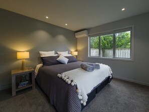 A bedroom with a large bed, two bedside lamps, a window with a view of greenery, and a built-in air conditioning unit.