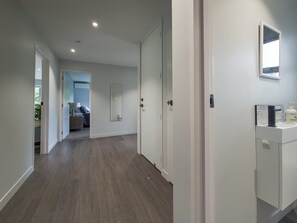 A hallway with a wooden floor, white walls, and a door leading to a room with a bed and a window. - Beachside Bliss - Two Bedroom Apartment (Napier)