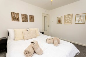 Ein ordentlich bezogenes Bett mit weißer Bettwäsche und gerollten Handtüchern, ein Nachttisch und drei an der Wand befestigte Kunstwerke.