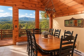 Una sala da pranzo in legno con un tavolo grande e sedie, un lampadario e vista sulle montagne attraverso ampie finestre.