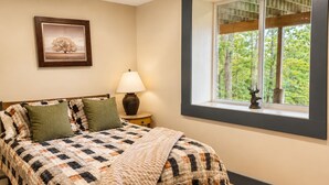 Quarto com cama, mesa de cabeceira, abajur, janela com vista para árvores e um quadro na parede.