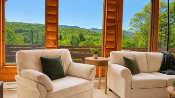 Una habitación con dos sofás color beige, una mesita redonda y vistas a un paisaje boscoso a través de amplios ventanales.