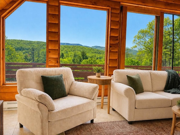 A room with two beige sofas, a small round table, and a view of a forested landscape through large windows.