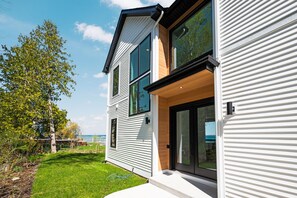 Ein modernes, zweistöckiges Haus mit großem Glasflureingang, einem sichtbaren Außenbereich mit Rasen und einem Baum im Hintergrund.