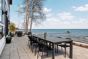 An outdoor patio with a table and chairs, overlooking a body of water with a boat. - Luxury Home offering 100 feet of private, sandy beachfront on beautiful West Bay |  West Bay Haven (Traverse City)