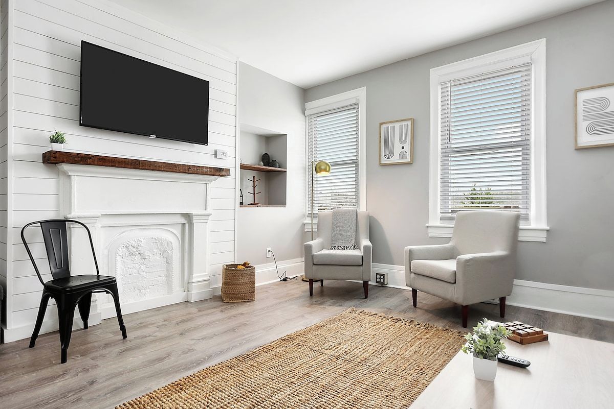 A living room with a fireplace, two armchairs, a television, and a large window with blinds.