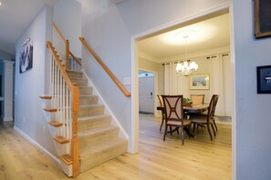 Una escalera con barandillas de madera y peldaños alfombrados en tono beige que conduce a un comedor con una araña y sillas de madera.