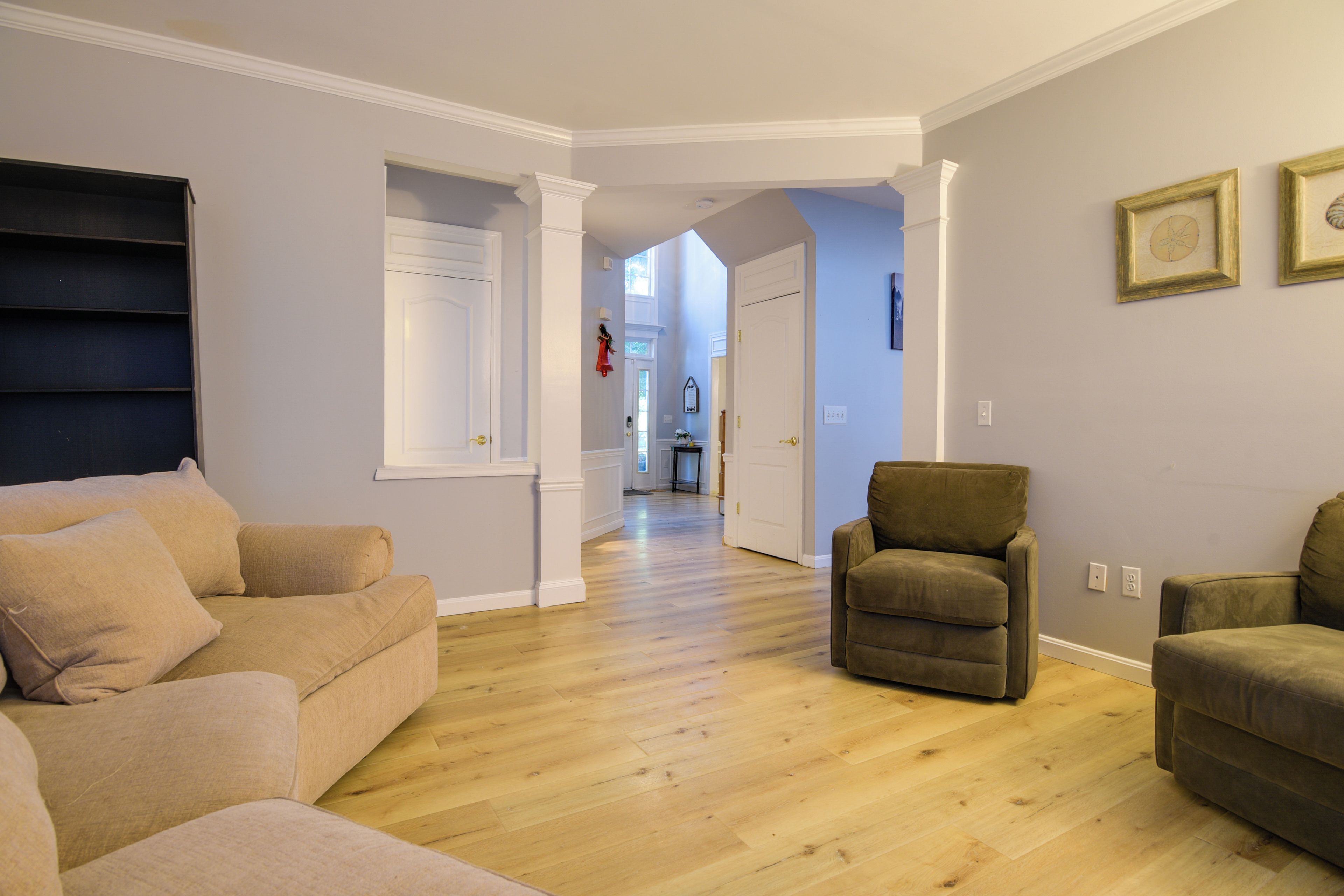 A spacious living room with a wooden floor, a beige sofa, a brown armchair, and a hallway leading to another room.