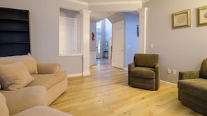 A spacious living room with a wooden floor, a beige sofa, a brown armchair, and a hallway leading to another room.
