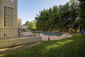 Un patio trasero con un área pavimentada, una valla metálica y un árbol grande.