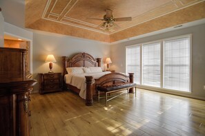 Un dormitorio con cama de madera, dos mesitas de noche con lámparas, un ventilador de techo y ventanas grandes con persianas.