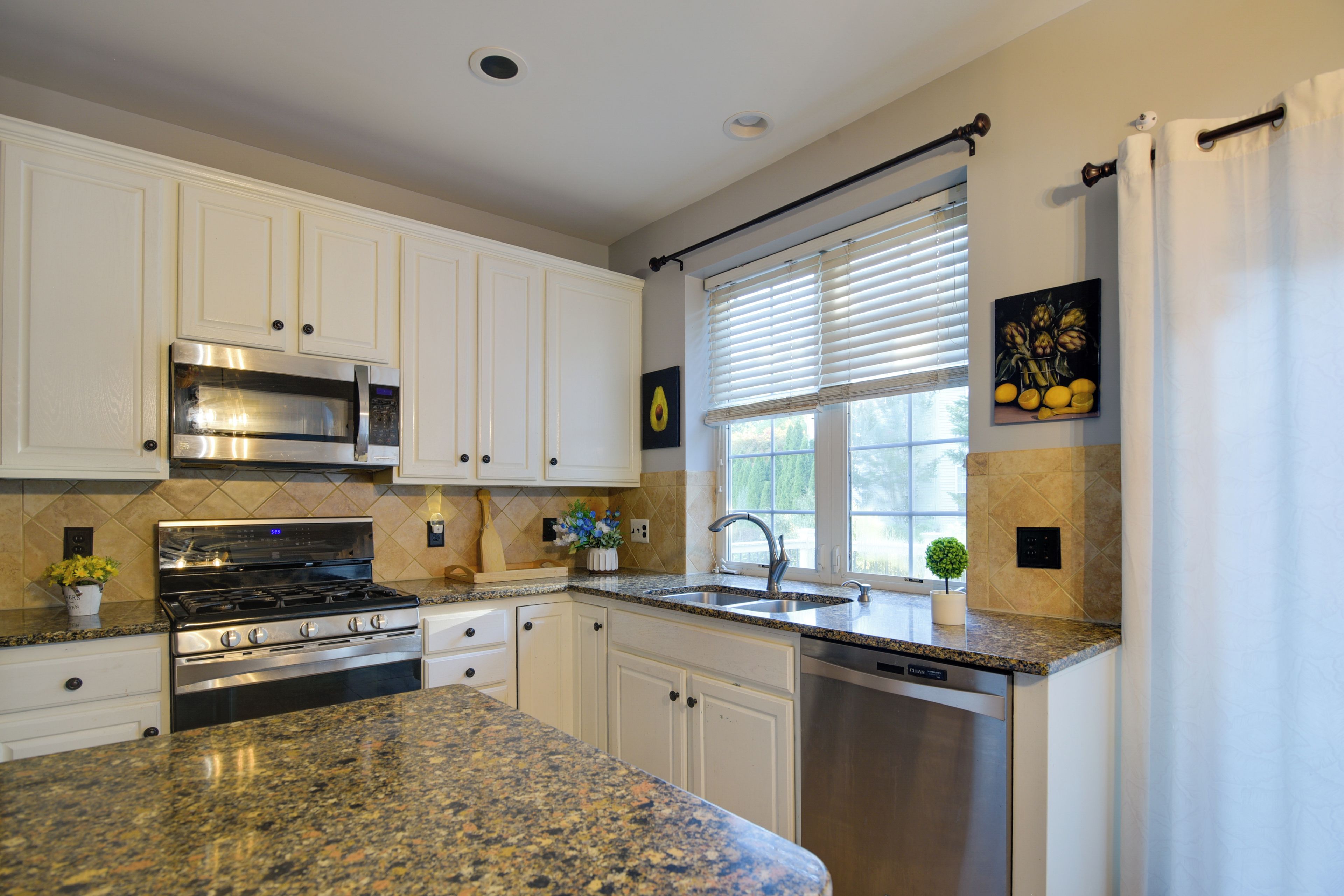 A modern kitchen with white cabinets, stainless steel appliances, granite countertops, and a window with blinds.