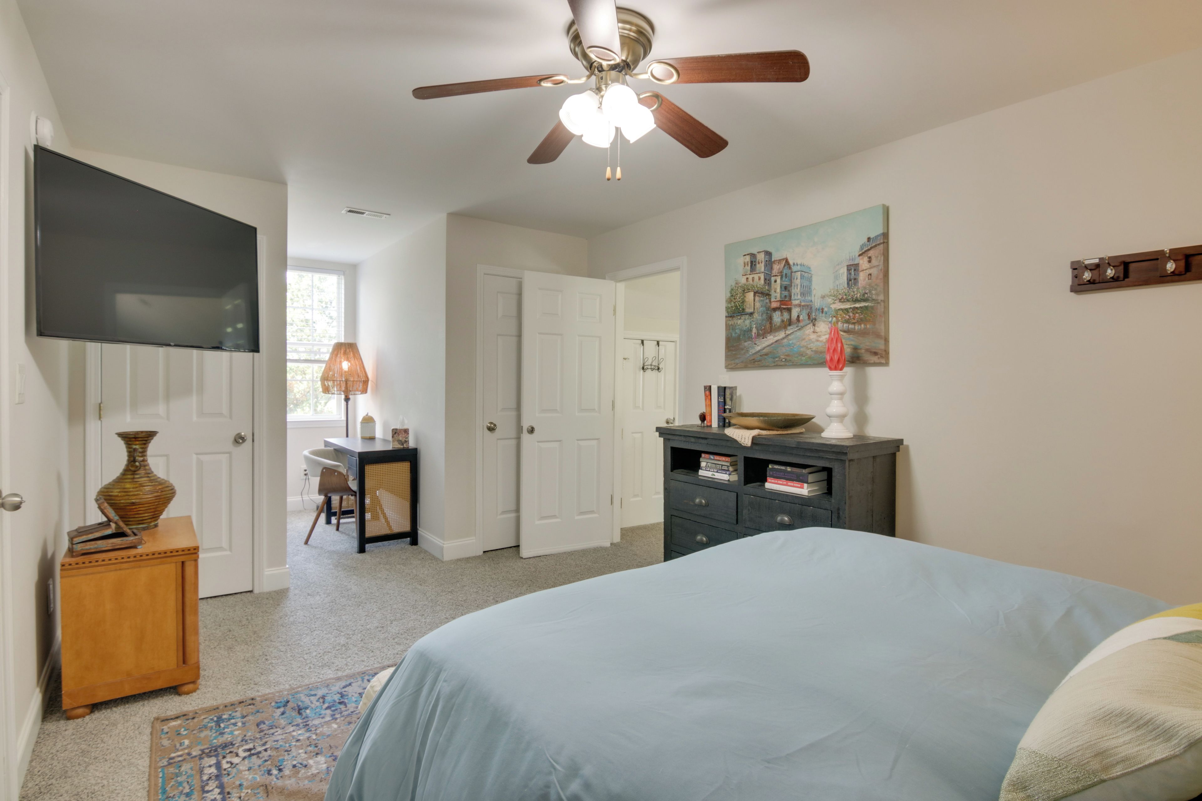 A bedroom with a bed, a ceiling fan, a desk with a lamp, a bookshelf, and a painting on the wall.
