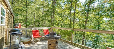 A wooden deck with a stone fire pit, red chairs, and a grill.
