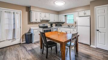 A compact kitchen with white cabinets, a wooden dining table, and a trash can.